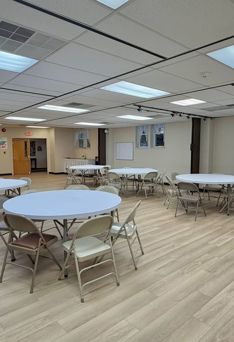 The renovated fellowship hall at a Clark congregation, seen down the length of the room. Round banquet tables and stacking chairs stand on a new light plank floor beneath a new suspended ceiling.