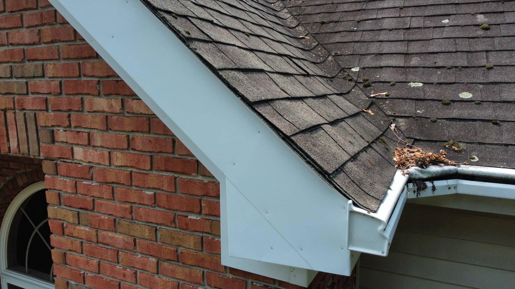 Before, detail: cupped and granule-worn shingles with moss growth and leaf debris packed in the gutter at the brick gable