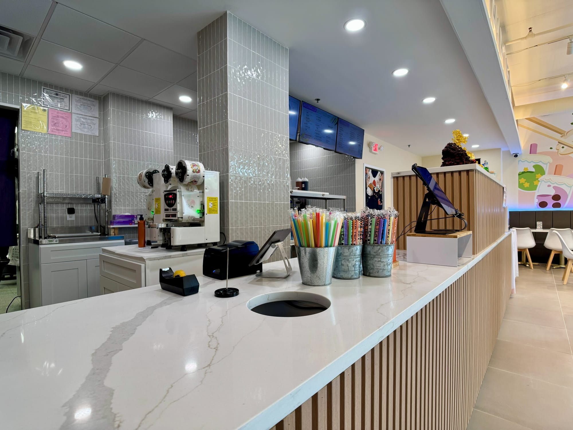 Service counter in quartz with slatted-oak front, sealing machine and straw caddies, menu screens above